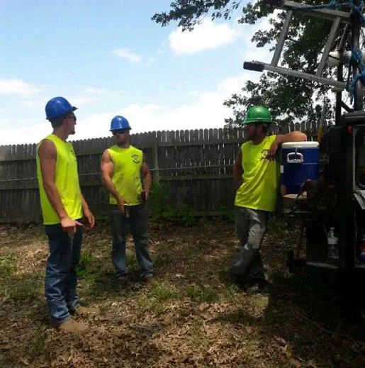Three construction workers in neon yellow shirts and hard hats stand near a truck, engaged in conversation with trees and a wooden fence in the background.