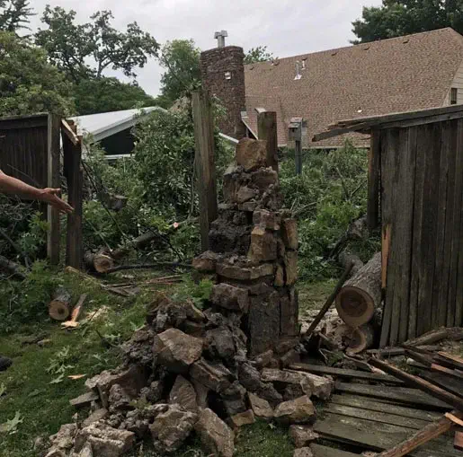 A wooden fence and stone pillar have collapsed, with large tree branches scattered around the yard. A person's arm is visible on the left, gesturing toward the damage. Houses and trees are in the background.