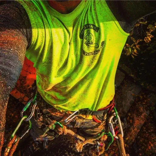 Person wearing a bright yellow shirt with a logo, harness, and ropes, appears to be engaged in a climbing activity. Their clothes and gear are covered in dirt.