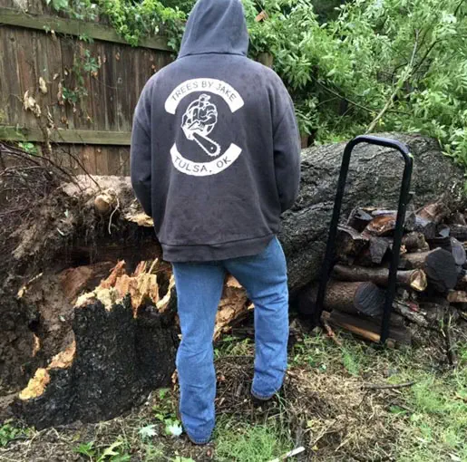 A person in a "Trees by Jake - Tulsa, OK" hoodie stands beside an uprooted tree and a stack of firewood in a wooded area.