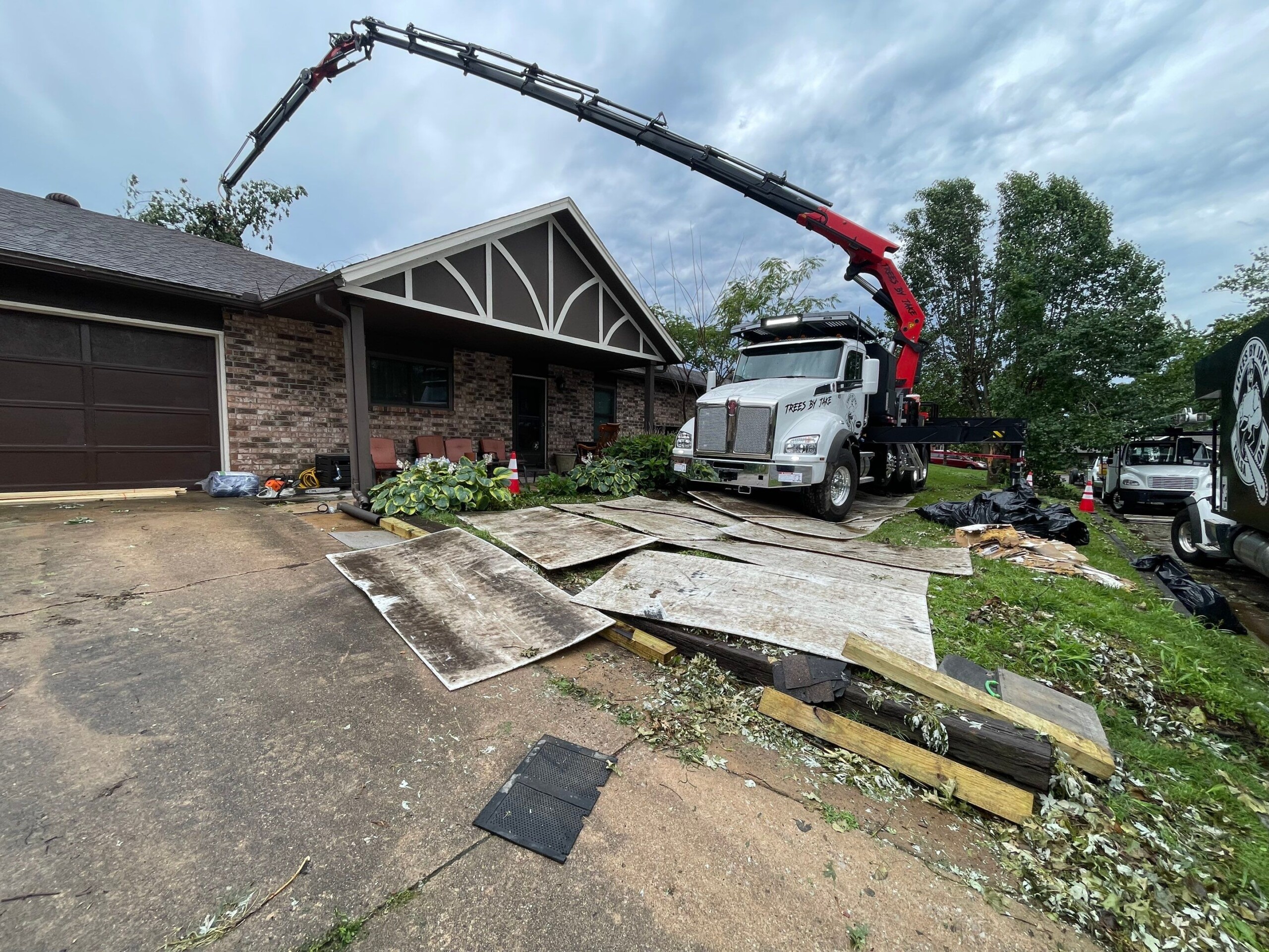 A truck with a large crane arm parked in front of a house with its roof partially torn off, debris and removed roof panels scattered on the ground. Tree branches and workers are visible in the background.