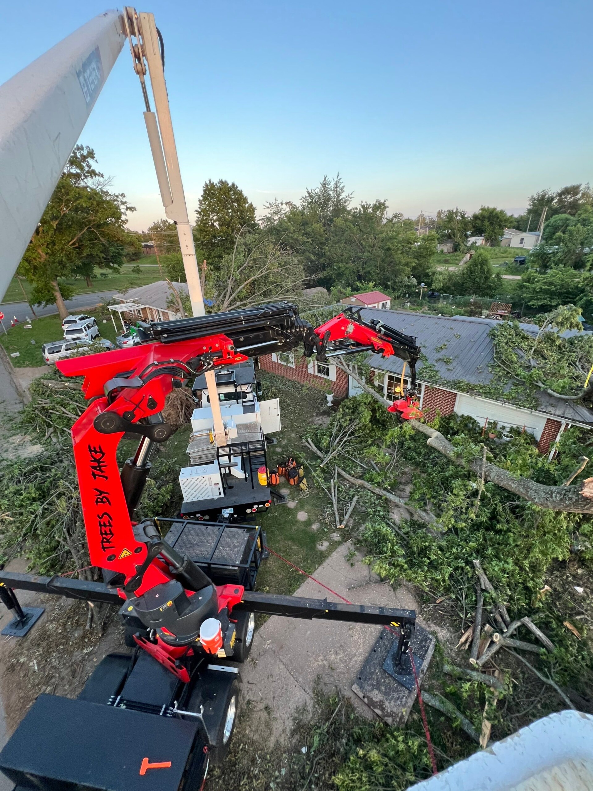A red crane truck with extended arm cuts and removes large tree branches near a residential house, with fallen branches scattered on the ground.