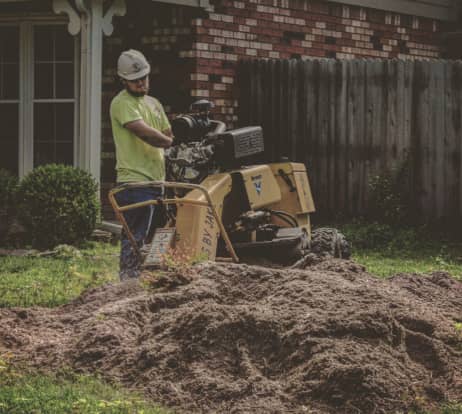 A worker in a hard hat operates a stump grinder next to a large pile of mulch in a residential backyard.