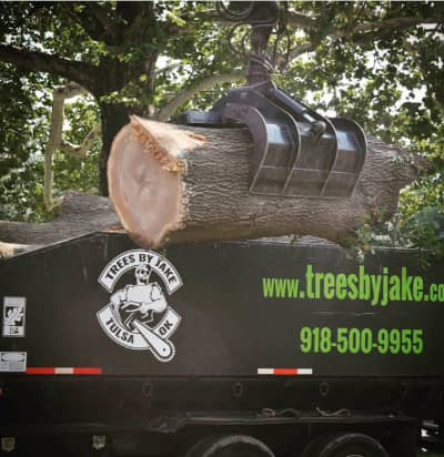 Large log being lifted onto a truck with "Trees by Jake Tulsa OK" logo, website, and phone number visible; tree branches in the background.