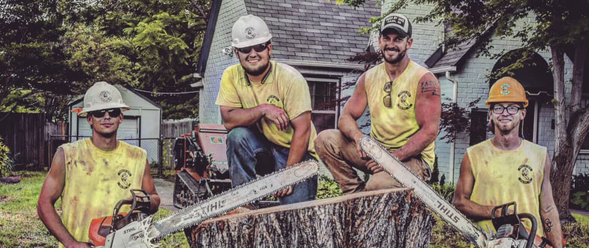 Four workers in yellow shirts and hard hats pose with chainsaws around a large tree stump in front of a house.