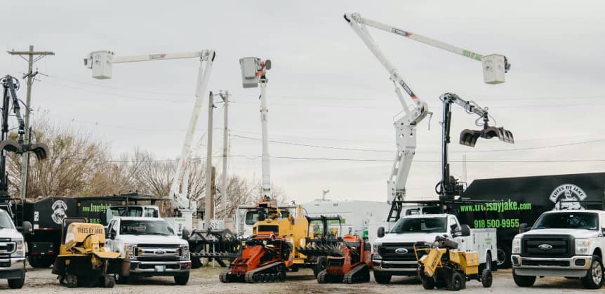 A row of utility trucks and machinery, including bucket trucks and stump grinders, parked on a gravel lot with trees and a company truck in the background.
